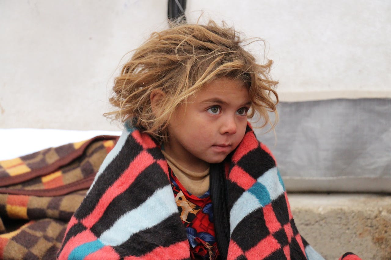 Een klein kind zit buiten onder een deken in een vluchtelingenkamp