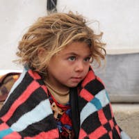 Een klein kind zit buiten onder een deken in een vluchtelingenkamp