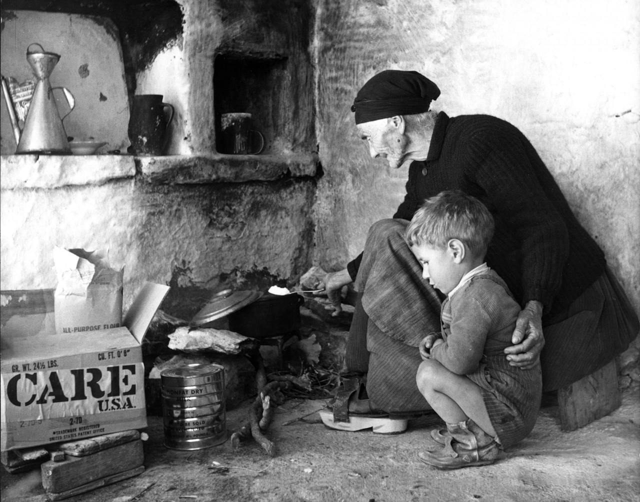 Historische zwart-witfoto: een oude vrouw zorgt in haar simpele keukentje voor haar wees geworden kleinzoon door een maaltijd voor hem te koken met de producten uit het CARE-pakket na de Tweede Wereldoorlog in Griekenland