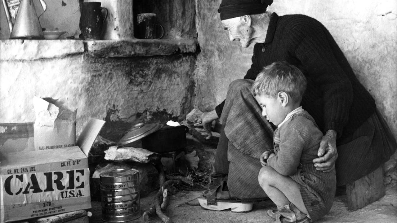 Historische zwart-witfoto: een oude vrouw zorgt in haar simpele keukentje voor haar wees geworden kleinzoon door een maaltijd voor hem te koken met de producten uit het CARE-pakket na de Tweede Wereldoorlog in Griekenland